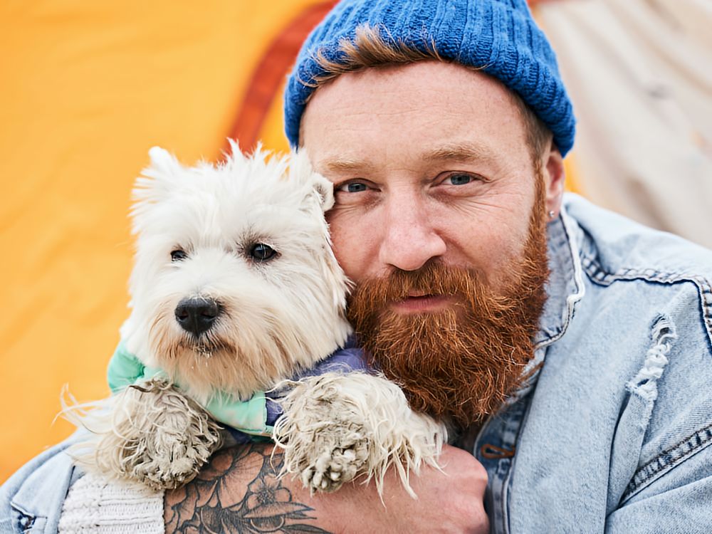 Pet Adoption Man with a knit hat and beard holding a white terrier up to his face in an endearing snuggle