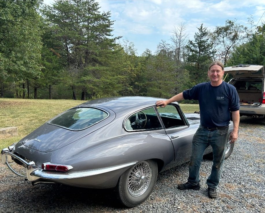 A smiling man stands on a gravel driveway beside a silver vintage sports car, resting one hand on the roof. Behind him is a grassy yard bordered by trees under a partly cloudy sky.